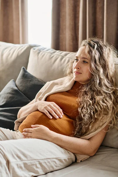 妊娠中の女性とともに長いですブロンド波毛を保ちます手上の彼女の腹中でリラックスしながら柔らかいソファと見ますテレビ レジャー — ストック写真