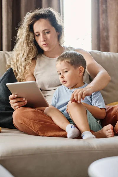 年轻而严肃的金发女人 带着平板电脑和她可爱的小儿子坐在沙发上看网上儿童电影或卡通片 — 图库照片