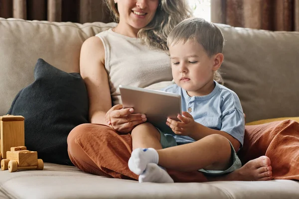 认真的小孩穿着便服 一边坐在沙发上 一边看卡通片 一边看着妈妈拿着的平板电脑屏幕 — 图库照片