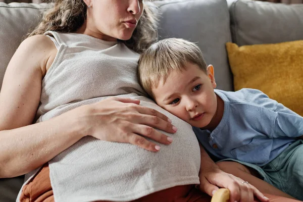 好奇心旺盛な男の子彼の妊娠中の母親の腹の上に頭を維持しながら ソファに座っている間 子宮内で胎児のハートビートを聞いて — ストック写真