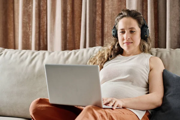 若いです妊娠中の女性とともにラップトップ上の彼女の膝見ますオンラインビデオまたは通信誰かと一緒にリラックスしながらソファ — ストック写真
