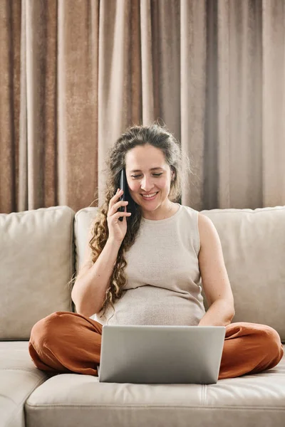 若いです妊娠中の女性とともに長いブロンド波状毛注文商品によって携帯電話で座っている間ソファとラップトップ画面を見ます — ストック写真