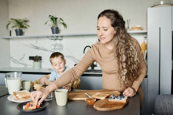 若いです妊娠中の女性Brendingオーバーキッチンテーブルながら準備サンドイッチとともにブルーベリーで朝食のために彼女の小さな息子 — ストック写真