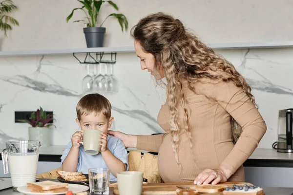 若いです妊娠中の女性立ち隣に彼女の小さな息子とともにマグカップ飲料牛乳や紅茶ながら 朝食のための自家製サンドイッチを持って — ストック写真