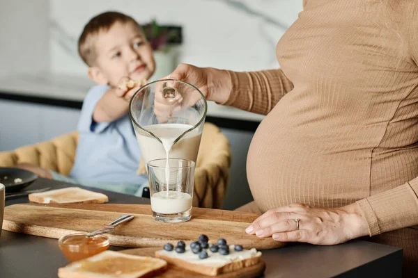 若いです妊娠中の女性の閉じ込めガラスに新鮮な牛乳を注ぐのために彼女の可愛いです息子のために座っていますアームチェアでキッチンテーブル — ストック写真