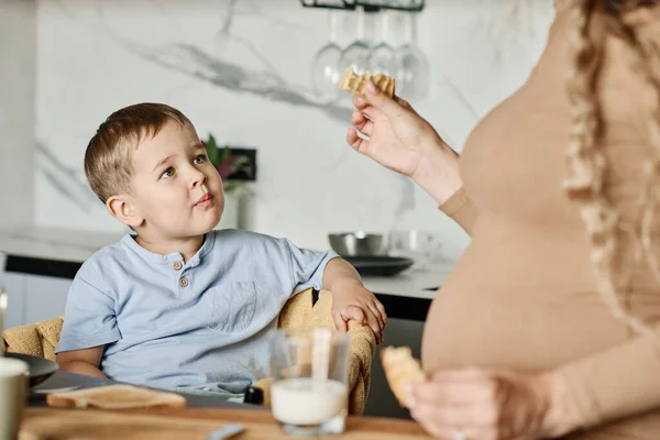 可爱的小男孩一边在厨房桌子边吃早饭 一边手里拿着一块自制饼干看着他怀孕的妈妈 — 图库照片