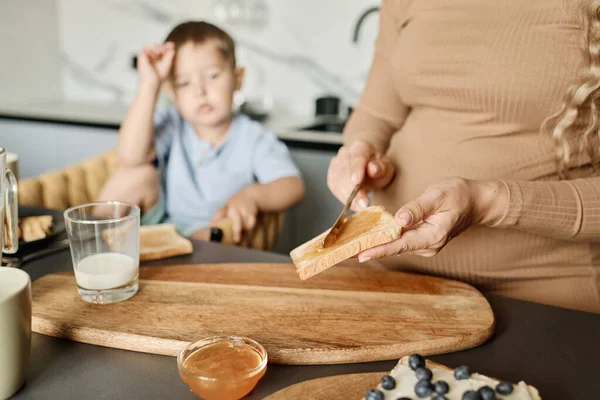 朝食のための彼女の小さな息子のためのスナックを準備しながら 若い妊婦の手は 小麦パンのスライスに蜂蜜やジャムを広げる — ストック写真
