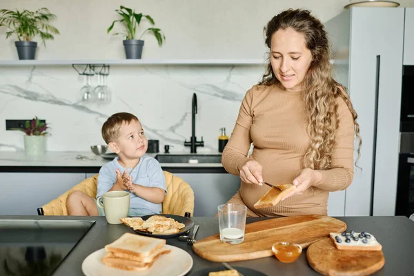 台所のテーブルに立っている若い妊婦は 朝食を準備しながら 小麦パンのスライスに蜂蜜やジャムを広める — ストック写真