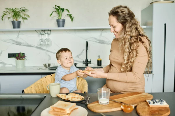年轻而细心的母亲准备着蜂蜜或果酱三明治 然后把它递给坐在厨房桌子旁边的快乐的小儿子 — 图库照片