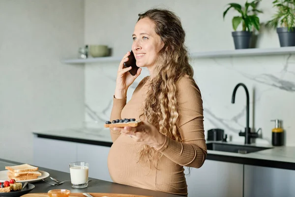 若い陽気妊娠中の女性とサンドイッチは 朝食中にキッチンテーブルに立っている間 スマートフォン上で通信 — ストック写真