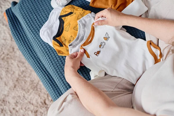上图拍摄的年轻孕妇坐在床上 在几件衣服前 为宝宝选择新的T恤衫 — 图库照片