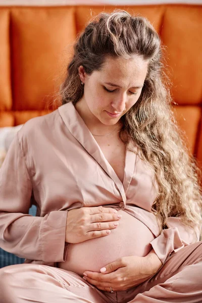 妊娠中の女性でピンクシルクパジャマ彼女の腹を見て それに触れながらベッドに座って 彼女の将来の赤ちゃんと通信 — ストック写真
