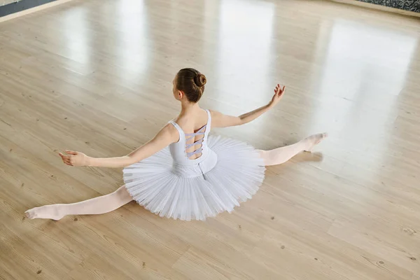 身着白色燕尾裙的优雅芭蕾舞演员坐在宽敞的教室地板上 从背角上方伸出胳膊和腿 — 图库照片