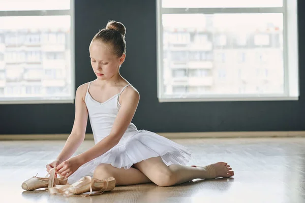 穿着白色晚礼服的年轻优雅的芭蕾舞演员坐在教室的地板上 解开米色针尖鞋的丝带 — 图库照片