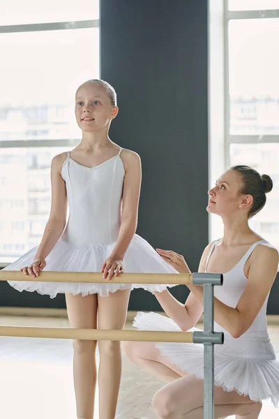 Young ballet teacher looking at youthful learner in white tutu standing by bar in front of mirror and practicing exercise at lesson