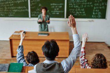 Bir kaç genç öğrencinin, öğretmenlerinin önünde otururken ellerini havada tutarak yeni bir konuyu açıklamaya çalışmalarına dikkat edin.