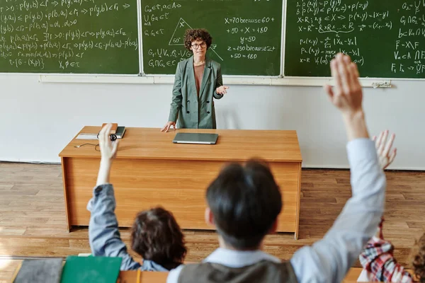 自信的老师站在黑板旁边 一边看着一群高举着手的学生 一边拿着公式和方程式 — 图库照片