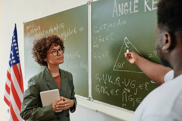 年轻而严肃的几何学女教师 听着非洲裔美国学生用粉笔指着黑板上的三角形 — 图库照片