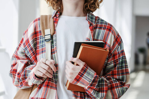 Hands of youthful female student in white t-shirt and checkered shirt holding book and copybook while standing in front of camera