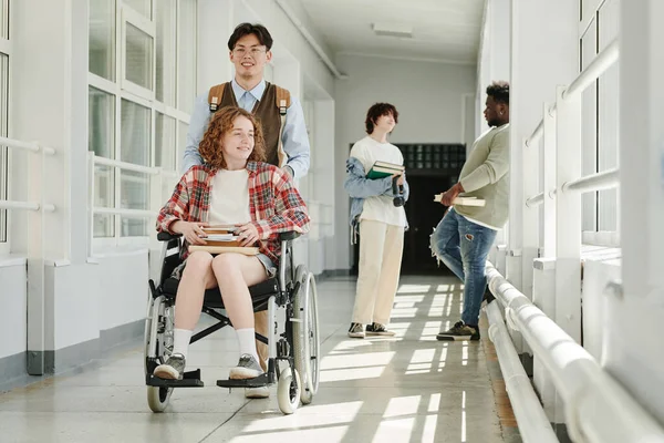快乐的亚洲少年把他的女友推上轮椅 沿着大学走廊向前走 然后走进教室 图书馆或食堂 — 图库照片