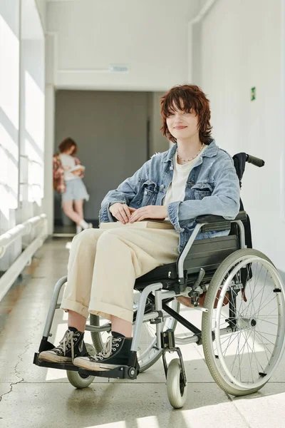 Happy teenage student with physical disability sitting in wheelchair in college corridor and looking at camera against her classmate