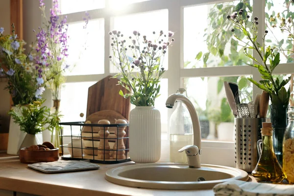 木や鋼の台所用品に囲まれた円形のシンクの上にベージュの金属タップや窓による花と花瓶 — ストック写真