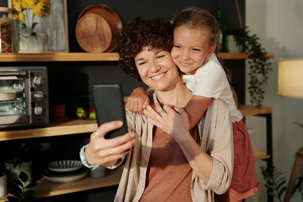 年轻的笑着的妈妈拿着智能手机 带着快乐的年轻女儿抱着自己 站在家里 — 图库照片