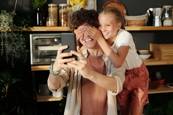 年轻可爱的女孩一边用智能手机捂住妈妈的眼睛 一边在用厨房用具的架子上自娱自乐 — 图库照片