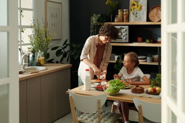 Genç esmer kadın, kızının yemek yediği mutfak masasının yanında dururken taze çavdar ekmeğine süt ürünleri serpiyor.