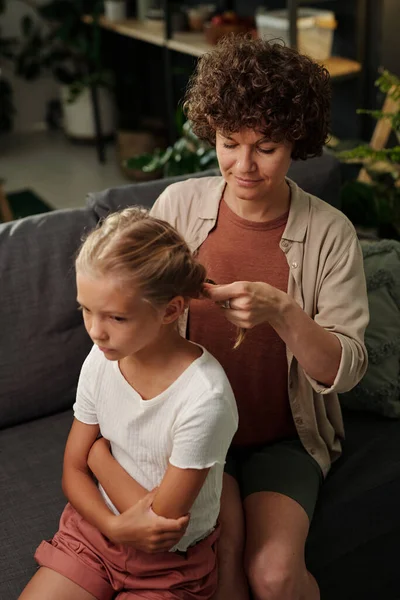 年轻的母亲穿着便服 在客厅里坐在沙发上 为她可爱的年轻女儿梳理着长长的金发 — 图库照片