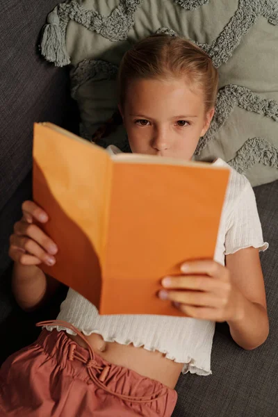 年轻可爱的女学生穿着便服 头枕在枕头上 在沙发上休息 阅读橙色封面的有趣书籍 — 图库照片