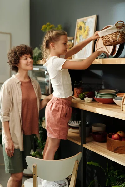 年轻女子站在她年幼的女儿身边 一边用厨房用具擦拭木架上的灰尘 一边帮助母亲 — 图库照片