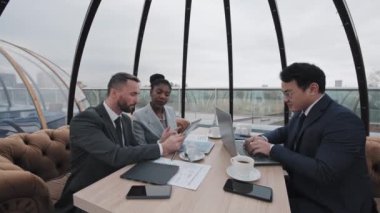 Asyalı ve Kafkasyalı erkek ve Afrikalı Amerikalı kadının resmi kıyafetlerle çatıdaki kafede oturup kahve içmeleri, belge ve bilgisayarlarla çalışmaları geniş açılı.