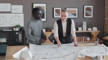 Gündüz vakti mimarlık bürosunda masa başında duran, mimari planlarla çalışan, konuşan Afrikalı genç erkek ve beyaz kadın.