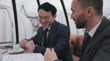 İki beyaz ve Asyalı resmi takım elbiseli adamın gündüz vakti açık hava restoranındaki masada oturup belgelerle konuşmalarının yan görüntüsü.