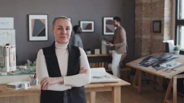 Kafası tıraşlı beyaz bir kadının orta boy portresi gündüz vakti mimarlık bürosunda çalışan meslektaşlarının önünde duruyor.