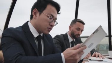 Tablet bilgisayar kullanarak resmi giyinmiş genç Asyalı iş adamının alçak açılı görüntüsü. Kafkasyalı erkek arkadaşı gündüz vakti açık hava restoranında masada oturuyor.