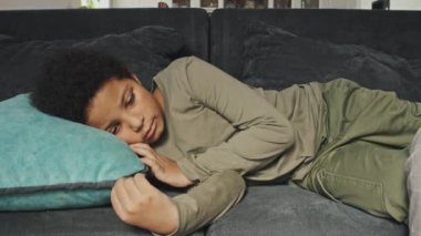 Orta uzunluktaki hasta Afrikalı Amerikalı çocuk evdeki kanepede yatıyor. Kırpılmış ebeveynin eli oğlunun alnına değiyor.