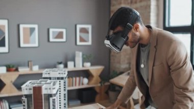 VR gözlüklü profesyonel çift ırklı erkek mimarın yan görüntüsü. Gündüz vakti ofisteki çok katlı apartman planına bakıyor.