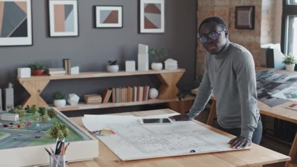 身着灰色套头衫的年轻黑人男子站在桌子旁边 头戴蓝图 白天在现代化的办公室里对着相机看 — 图库视频影像