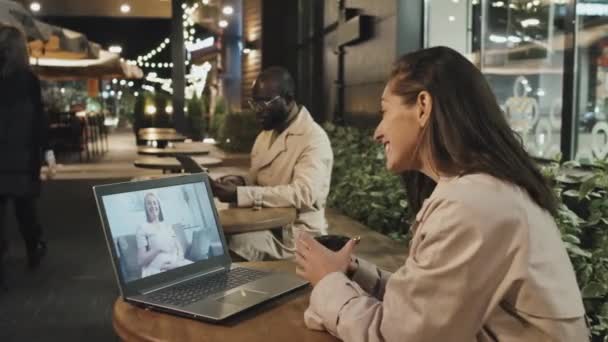 若いです白人女性座っていますカフェテーブル屋外で夜にチャットオンビデオコールとともに彼女の妹オンラップトップ 黒男使用してデジタルタブレット — ストック動画