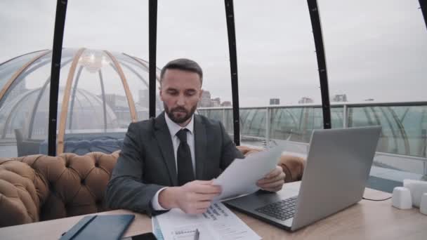 屋上のカフェでテーブルに座っているポータブルコンピュータと暗い灰色の正式なスーツで若い白人ビジネスマンの広い角度 文書を見て — ストック動画
