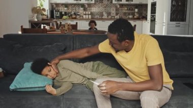 Orta boylu, kanepede oturan ve hasta oğlunun vücut ısısını ölçen Afrikalı Amerikalı bir adam.