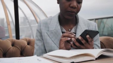 Gri takım elbiseli Afro-Amerikalı genç bir iş kadını çatıdaki restoranda oturmuş, akıllı telefon kullanıyor, not alıyor.
