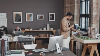 Orta boylu, çift ırklı erkek mimarın gündüz ofiste yürümesi, akıllı telefondan video araması, apartman planlarını göstermesi.
