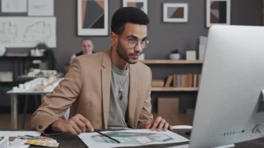 Genç, çift ırklı erkek tasarımcının belden yukarısında gündüz masa başında oturması, bilgisayar aracılığıyla video araması.