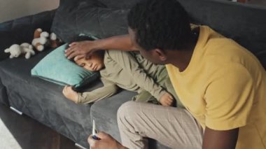 Kanepede oturan ve hasta oğlunun vücut ısısını ölçen Afro-Amerikan bir adamın orta boy yan görüşü.