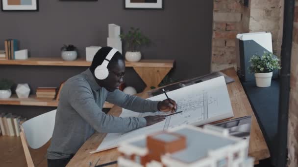昼間のオフィスの机に座って 青写真を描くヘッドフォンで若い黒人男性のサイドビューメディア — ストック動画