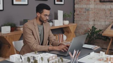 Orta boylu, zeki, çift ırklı bir mimar ofisinde oturuyor ve dizüstü bilgisayar kullanıyor.