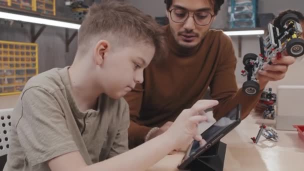 Tânăr Profesor Ajutându Elevul Construiască Model Robotic Privind Imaginea Tableta — Videoclip de stoc